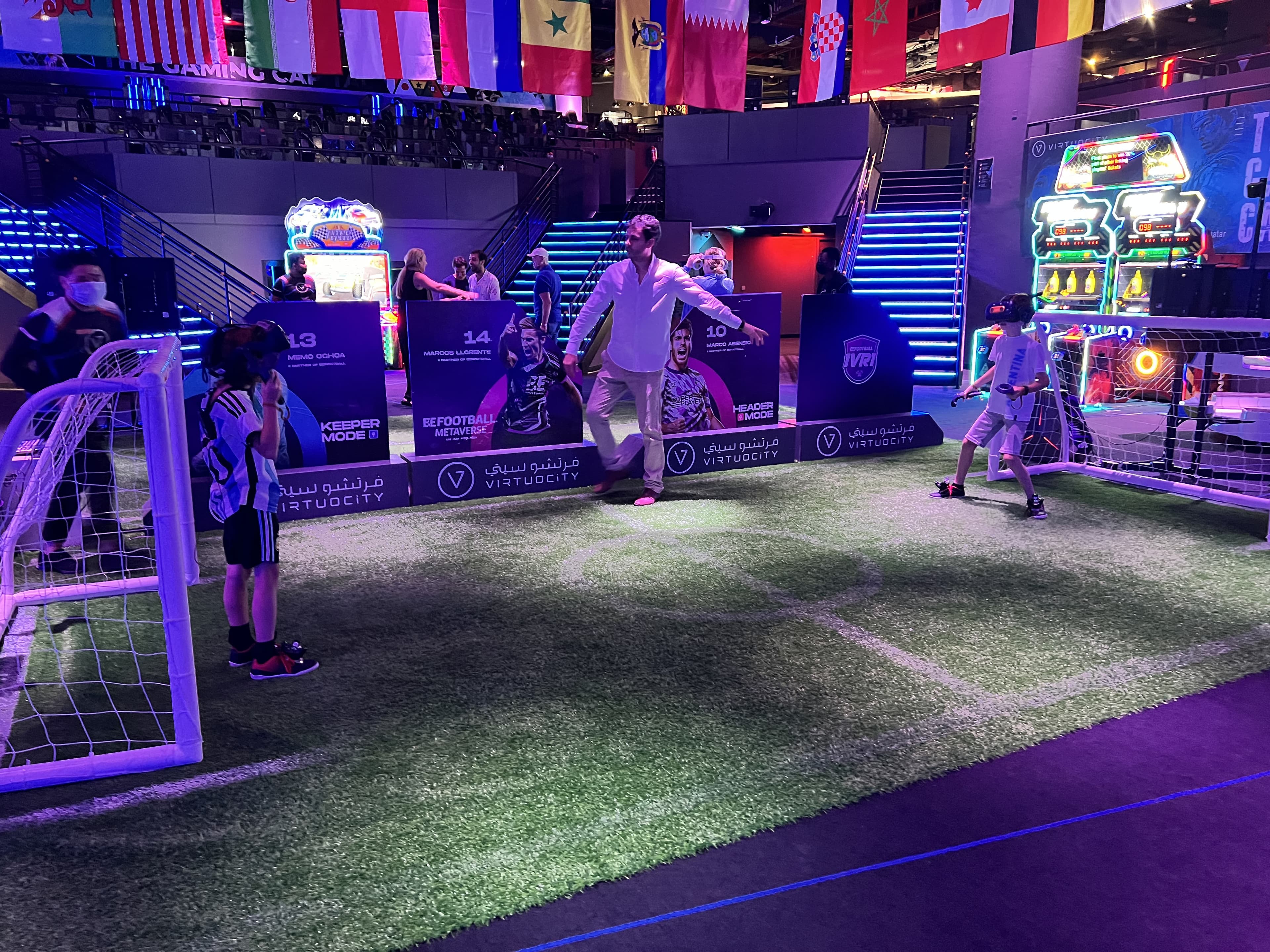 Children in VR headsets and an adult playing football on an indoor pitch in a neon-lit arcade hung with national flags